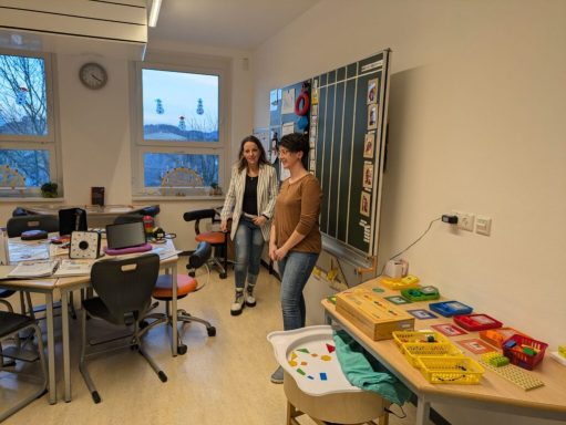 Zwei Frauen stehen in einem hellen Raum mit Tischen und Spielmaterialien.