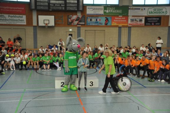 Ein Maskottchen steht neben Kindern in bunten T-Shirts in einer Sporthalle.
