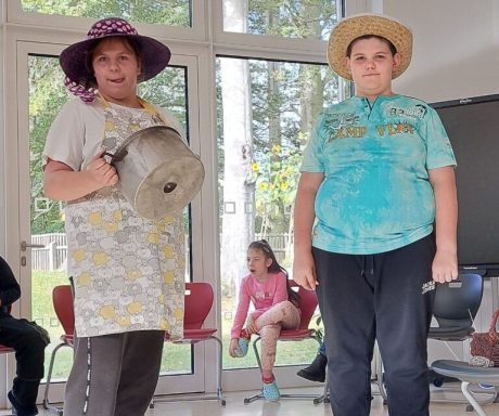 Zwei Personen in Sommerkleidung, eine mit Topf, die andere mit Hut, in einem Raum.