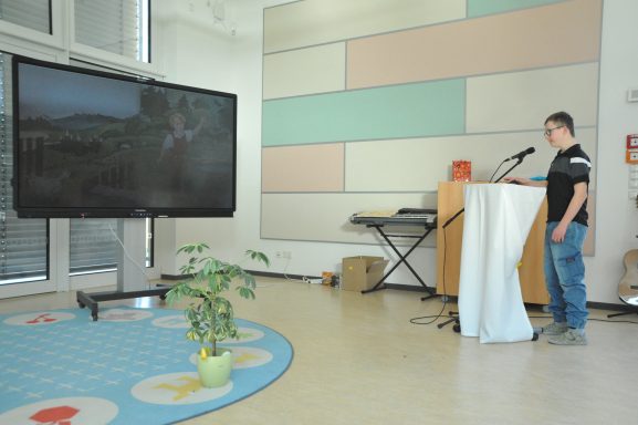 Mann steht an Mikrofon vor einem großen Bildschirm in einem hellen Raum mit Pflanzen.