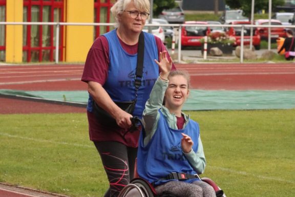 Eine Frau im Rollstuhl winkt, während eine Begleiterin sie schiebt. Beide tragen blaue Westen.