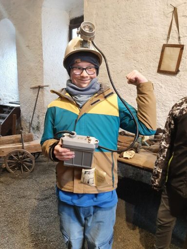 Person in Arbeitskleidung mit einem Helm und einer Grubenlampe hält ein Gerät in der Hand.