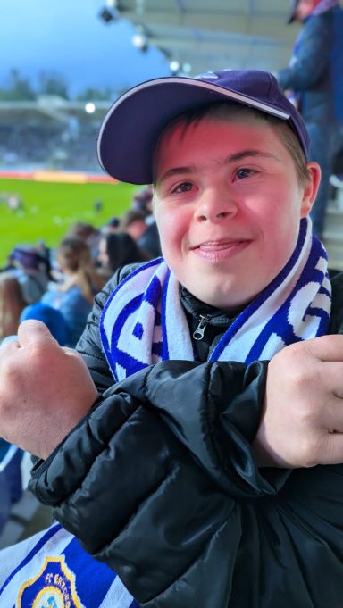 Junger Fan mit Schal und Mütze, jubelt im Stadion mit einer freundlichen Geste.