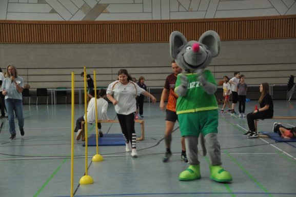 Ein Maskottchen in Mäusekostüm steht auf einem Sportplatz, während Teilnehmer um ihn herum laufen.