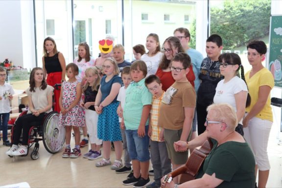 Gruppenszene mit Kindern und Erwachsenen, einige sitzen, andere stehen, lächeln.