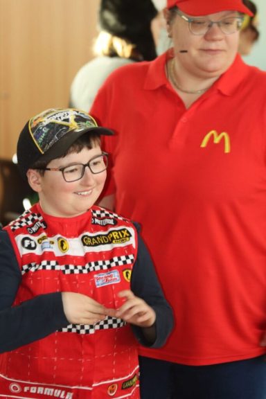 Ein Junge in einem Rennanzug mit McDonald's Symbol steht neben einer Frau mit rotem Shirt.