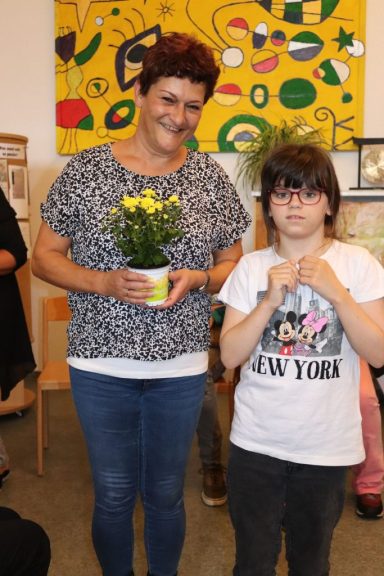Frau mit blühendem Topf in der Hand steht neben einem Kind. Beide lächeln.