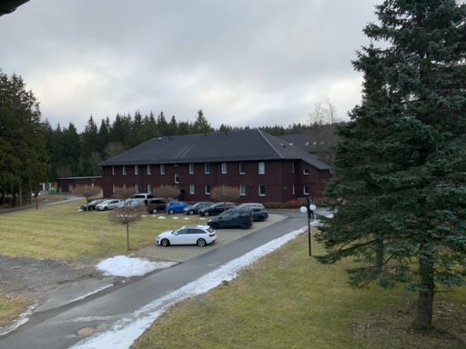 Rotes Gebäude mit Parkplätzen und Bäumen, umgeben von einer Wiesenlandschaft.