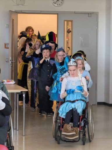 Kinder in bunten Kostümen betreten einen Raum, darunter ein Kind im Rollstuhl.