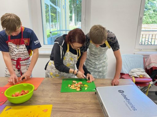 Drei Personen schneiden Gemüse in einer Küche, eine Person trägt eine Schürze.