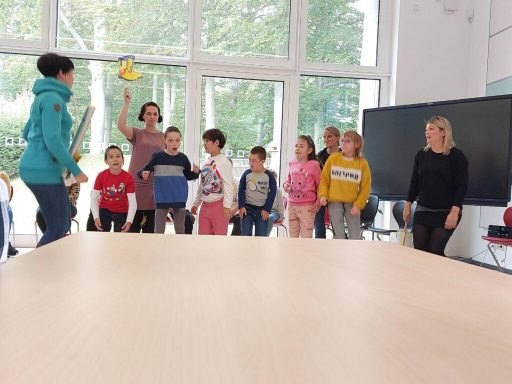 Kinder spielen in einem Raum mit großen Fenstern, eine Gruppe hat Spielzeug in der Hand.