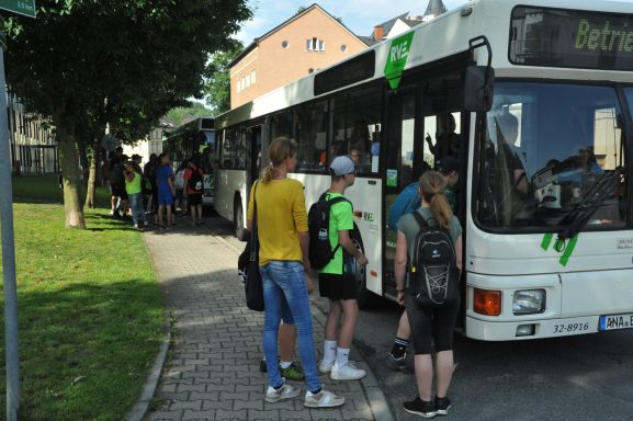 Bus mit Passagieren an einer Haltestelle, umgeben von Bäumen und Gebäuden.