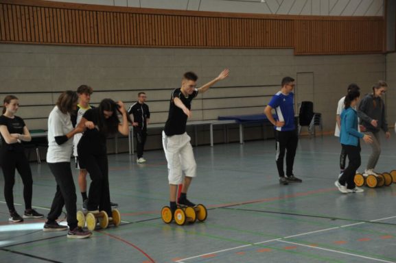 Gruppentraining in einer Sporthalle, Teilnehmer stehen auf Rollbrettern und balancieren.