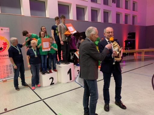 Zwei Männer sprechen auf einer Bühne, im Hintergrund stehen Kinder mit Preisen.