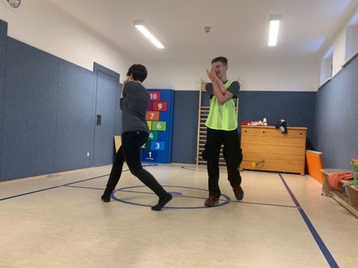 Zwei Personen üben in einem Raum mit Turnmatte und bunten Spielzeugen.