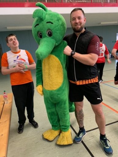 Zwei Personen in Sportkleidung, eine davon in einem grünen Maskottchen-Kostüm.