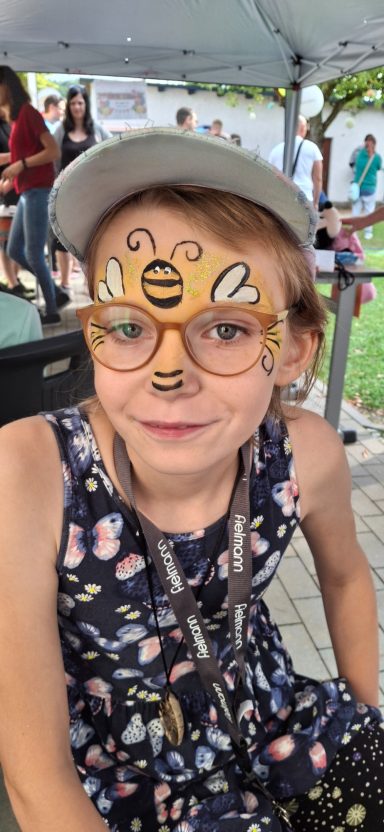 Ein Kind mit Bienen-Malerei im Gesicht und einer grauen Kappe, in einem Blumenkleid.