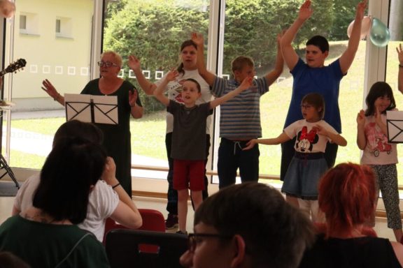 Gruppenaufführung mit Kindern und Erwachsenen, die in einer hellen Umgebung singen und tanzen.