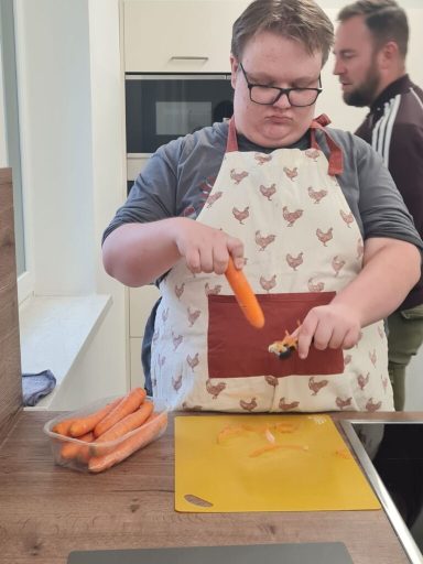 Junge schält Möhren in einer modernen Küche, während ein Mann im Hintergrund steht.