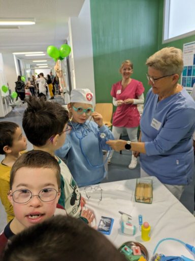 Ein Kind in Arztkleidung erhält Hilfe von einer Erwachsenen in einem Flur mit bunten Ballons.