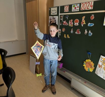 Ein Junge hält ein buntes Bild vor einer beschrifteten Tafel in einem Klassenraum.