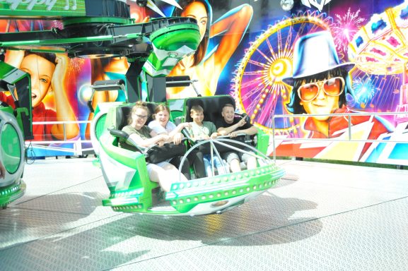 Vier Personen sitzen in einer Fahrgeschäft, umgeben von bunten Grafiken und Lichtern.