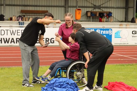 Eine Person im Rollstuhl erhält Unterstützung von drei Betreuern bei einer Sportveranstaltung.