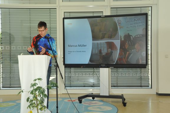 Eine Person präsentiert an einem Tisch vor einem großen Bildschirm in einem hellen Raum.