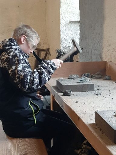Ein Junge bearbeitet einen Stein mit einem Hammer an einem Tisch in einem werkstattähnlichen Raum.