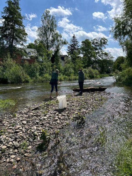 Zwei Personen stehen am Ufer eines Flusses, umgeben von Bäumen und klarem Himmel.