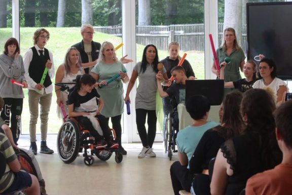Gruppe von Menschen, darunter Kinder und Erwachsene, stehen in einem Raum und halten bunte Stäbe.
