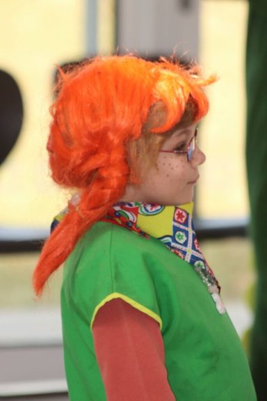 Kind mit orangefarbener Perücke und grüner Kleidung, seitliche Ansicht.