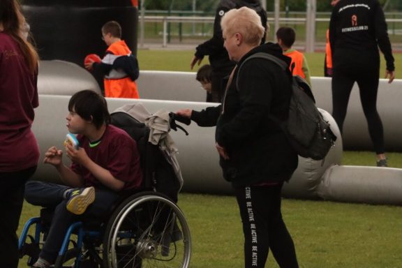 Junger Mann im Rollstuhl isst, während eine Frau mit schwarzer Jacke ihn beobachtet.