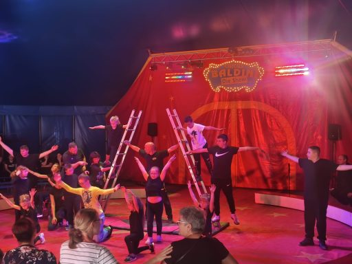 Zirkusaufführung mit Artisten in Kostümen vor einem bunten Zelt und Publikum.