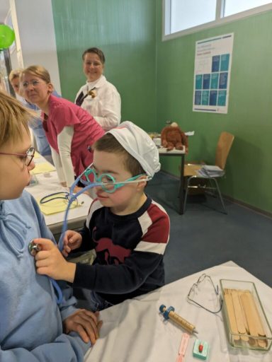 Kind trägt Brille und spielt Arzt, während Erwachsene im Hintergrund beobachten.