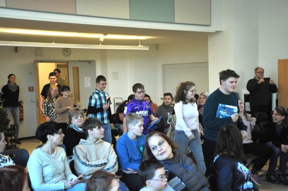 Gruppenszene mit Schülern in einem Raum, einige stehen, andere sitzen.