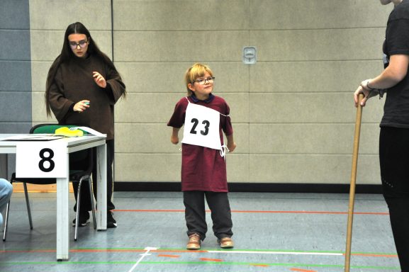 Ein blondes Kind mit der Nummer 23 steht in einem Raum, während eine Person mit einem Stock spricht.