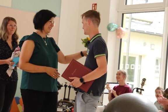 Eine Person überreicht einem jungen Mann ein Dokument, umgeben von Feiern.