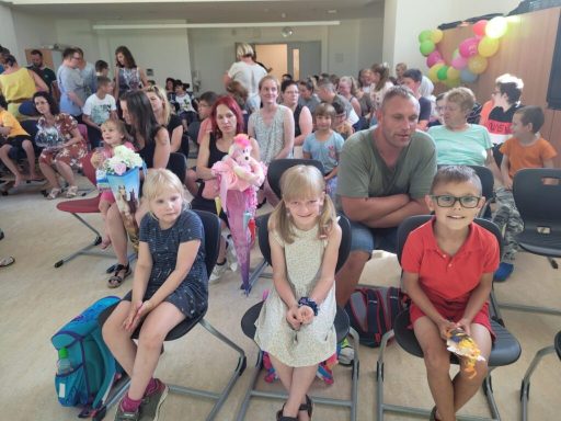 Gruppe von Kindern und Erwachsenen sitzt in einem Raum, bunte Ballons im Hintergrund.