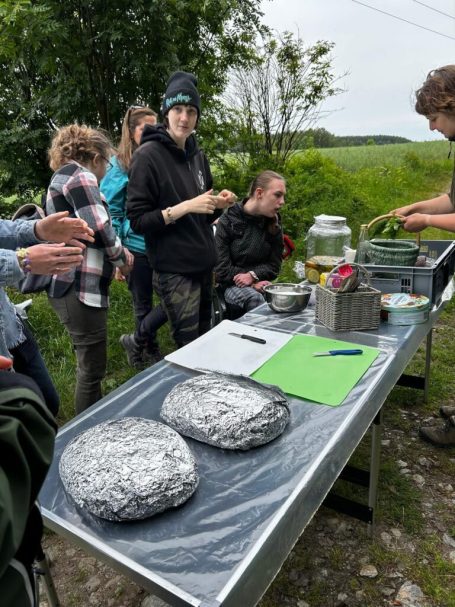 Menschen stehen um einen Tisch mit verschiedenen Utensilien und Speisen in einer grünen Landschaft.
