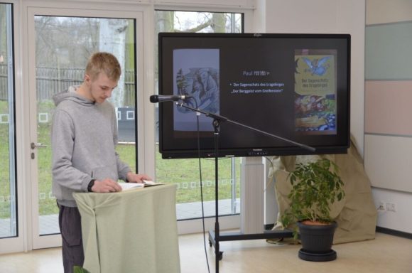 Junger Mann steht an einem Tisch, während er vor einem Bildschirm mit Präsentation spricht.