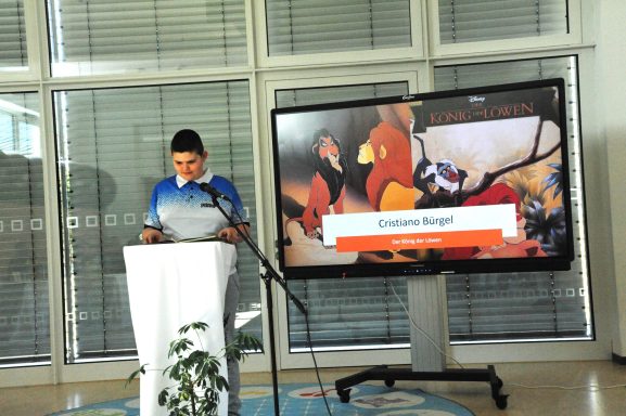 Junger Mann steht an einem Podium mit einem Bildschirm im Hintergrund.