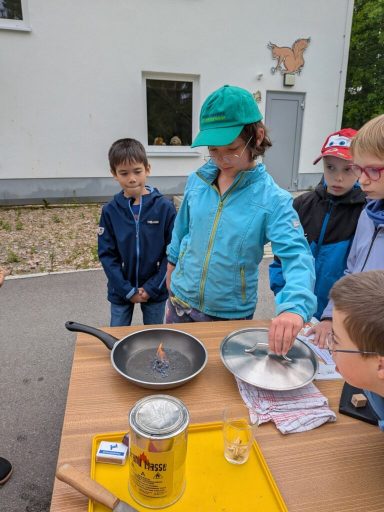 Kinder stehen um einen Tisch, während ein Mädchen eine Pfanne mit einem Deckel hält.