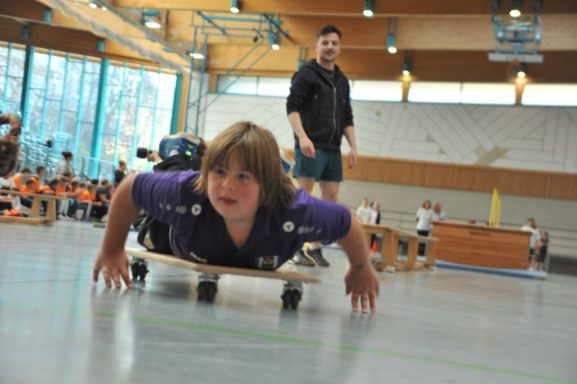 Ein Kind auf einem Skateboard, das am Boden liegt, ein Erwachsener beobachtet.