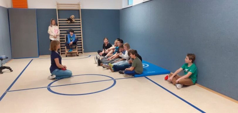 Eine Gruppe von Kindern sitzt auf dem Boden in einem Raum mit einer Trainerin.