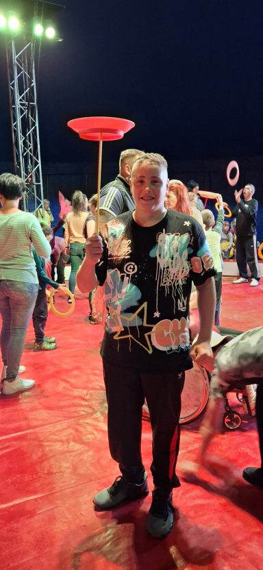 Ein Mann in einem T-Shirt mit Aufdrucken steht auf einem Zirkusboden mit anderen Leuten.