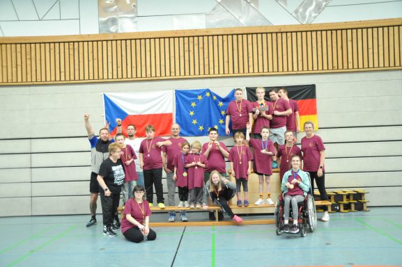 Gruppenselfie von Sportlern in lila T-Shirts vor Flaggen von Tschechien, Europa und Deutschland.