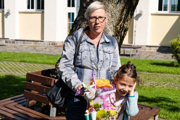 Eine Frau mit Brille steht neben einem Mädchen, das blühende Pflanzen hält.