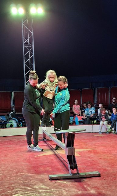 Zwei Personen unterstützen eine Akrobatin auf einem Balancebalken im Zirkus.