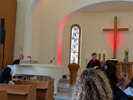 Ein Kirchenraum mit einem Pianisten und einem Redner vor einem Kreuz und bunten Fenstern.